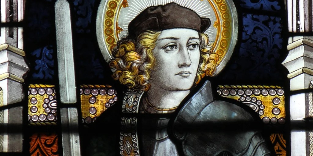 Stained glass depiction of a young knight with long, curly hair, wearing armor and a hat, holding a sword, against a decorative background.